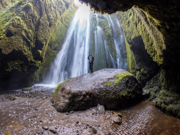 Person standing on a moss-covered rock inside a cave, looking toward a powerful waterfall illuminated from above.