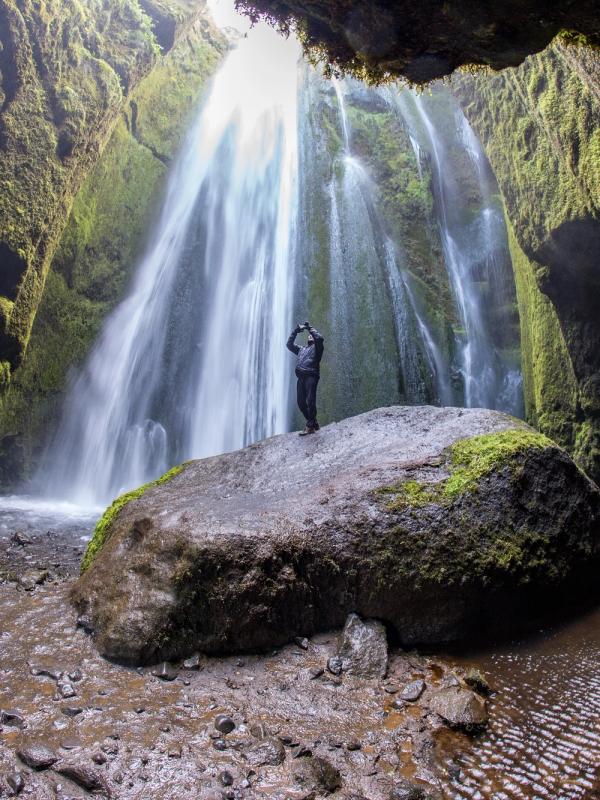 A person stands on a large mossy rock within a cave, looking up at a powerful waterfall descending from a bright opening above.