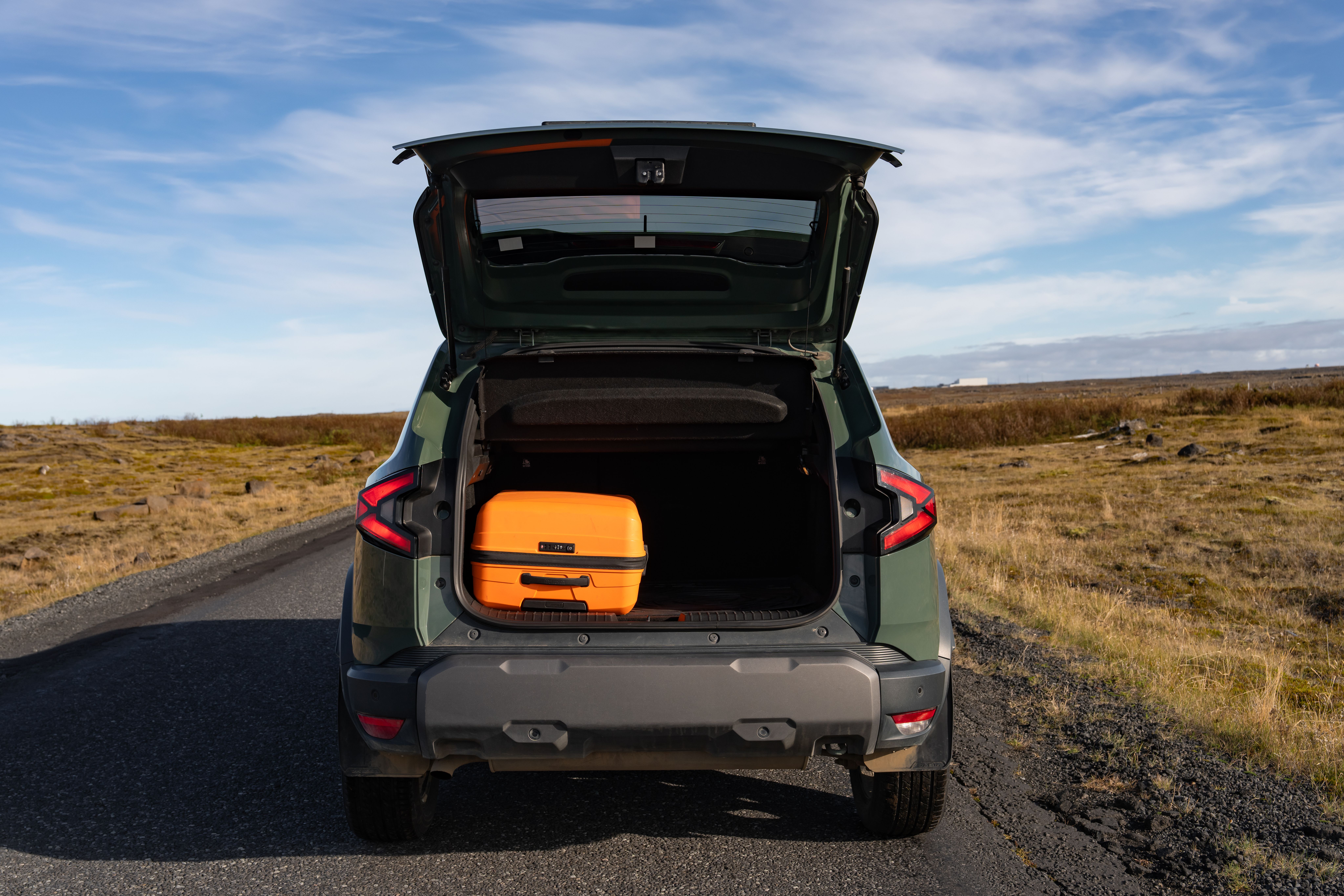 Rear of a dark green SUV with its trunk open, an orange suitcase inside, on a road in a dry landscape.