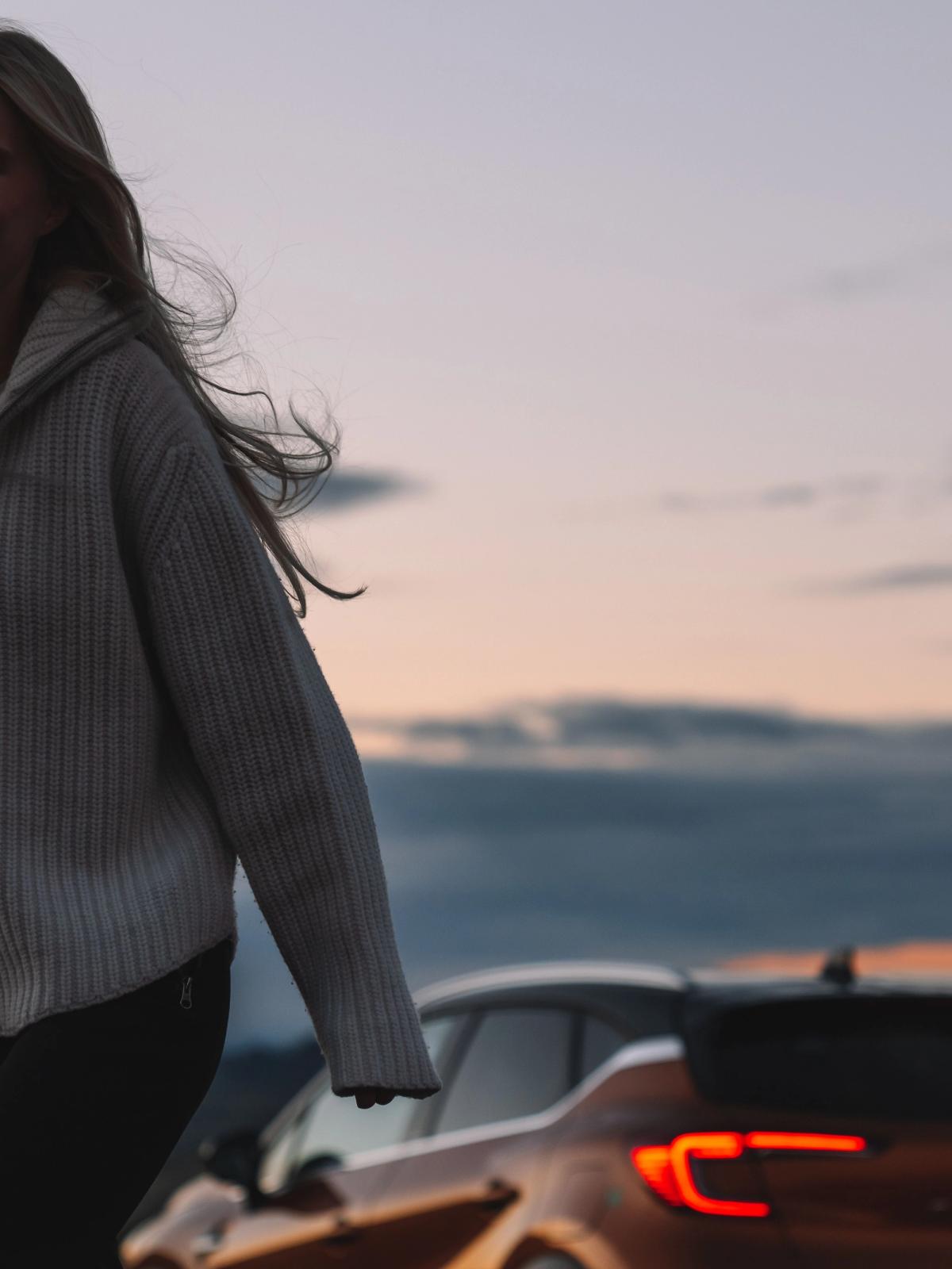 a woman in a sweater stands in front of two cars