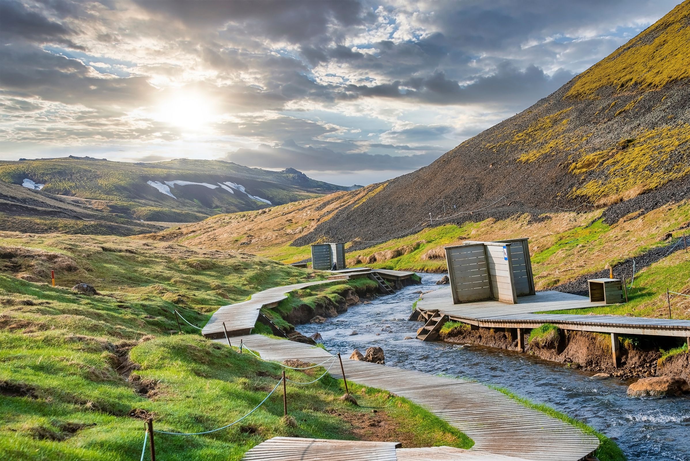 Wooden boardwalk and changing rooms beside a river in a sunny, green mountain valley.