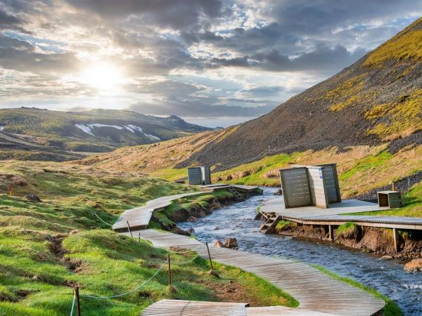 Wooden boardwalks and changing huts line a river flowing through a green mountain valley under a bright, cloudy sky.