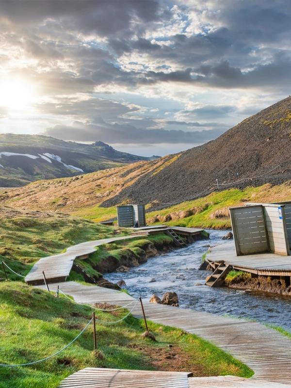 A wooden boardwalk and changing cabins follow a river through a green mountainous valley under a sunny, cloudy sky.