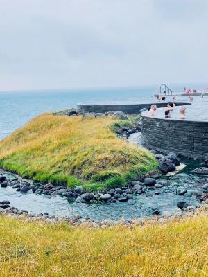 Outdoor thermal pool with people, overlooking the ocean and grassy hills with a stream.