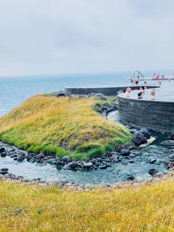 Outdoor thermal pool with people, overlooking the ocean and grassy hills with a stream.