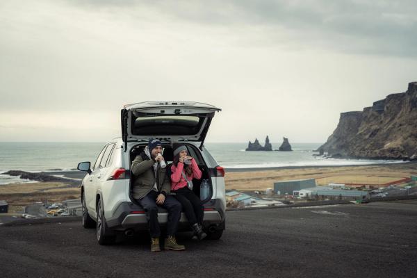 A couple sits in an open SUV trunk, drinking from mugs, overlooking a black sand beach, sea stacks, and cliffs.