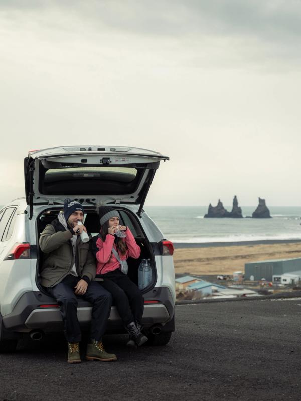 A couple sits in the open trunk of a white SUV, drinking, overlooking a black sand beach and sea stacks.