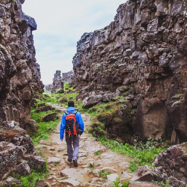 A hiker walks on a rocky trail between towering rock walls covered in green vegetation.