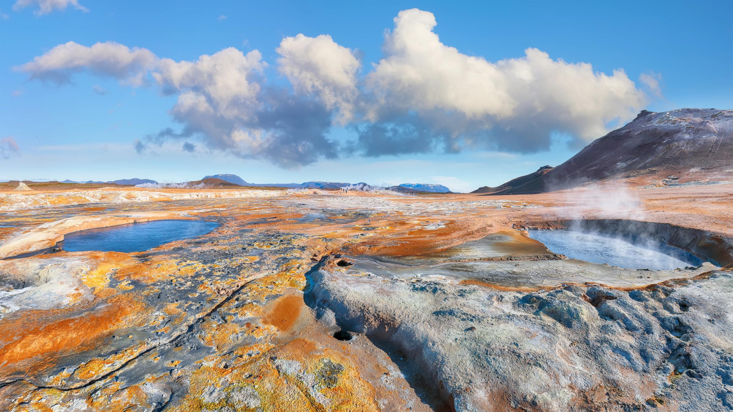 Un colorido paisaje geotérmico con manantiales de agua caliente humeantes, pozas azules y montañas lejanas bajo un cielo azul.