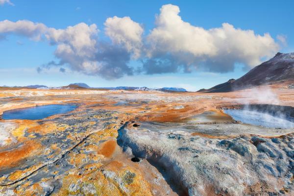 A colorful geothermal landscape with steaming hot springs, blue pools, and distant mountains under a blue sky.