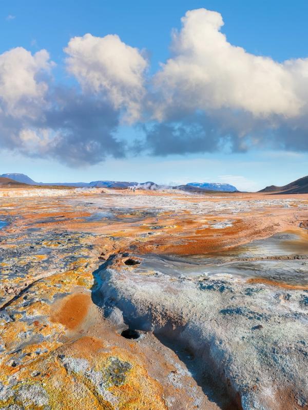 Vibrant orange and grey geothermal landscape with steaming pools and a still pond under a blue sky.