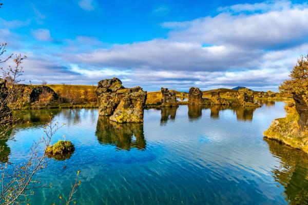 A blue lake dotted with rugged rock formations, surrounded by autumn trees under a cloudy sky.