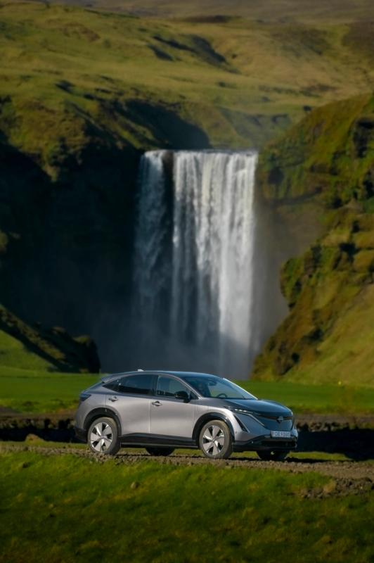 A silver crossover SUV on a dirt road in a green, hilly landscape with a large waterfall in the background.