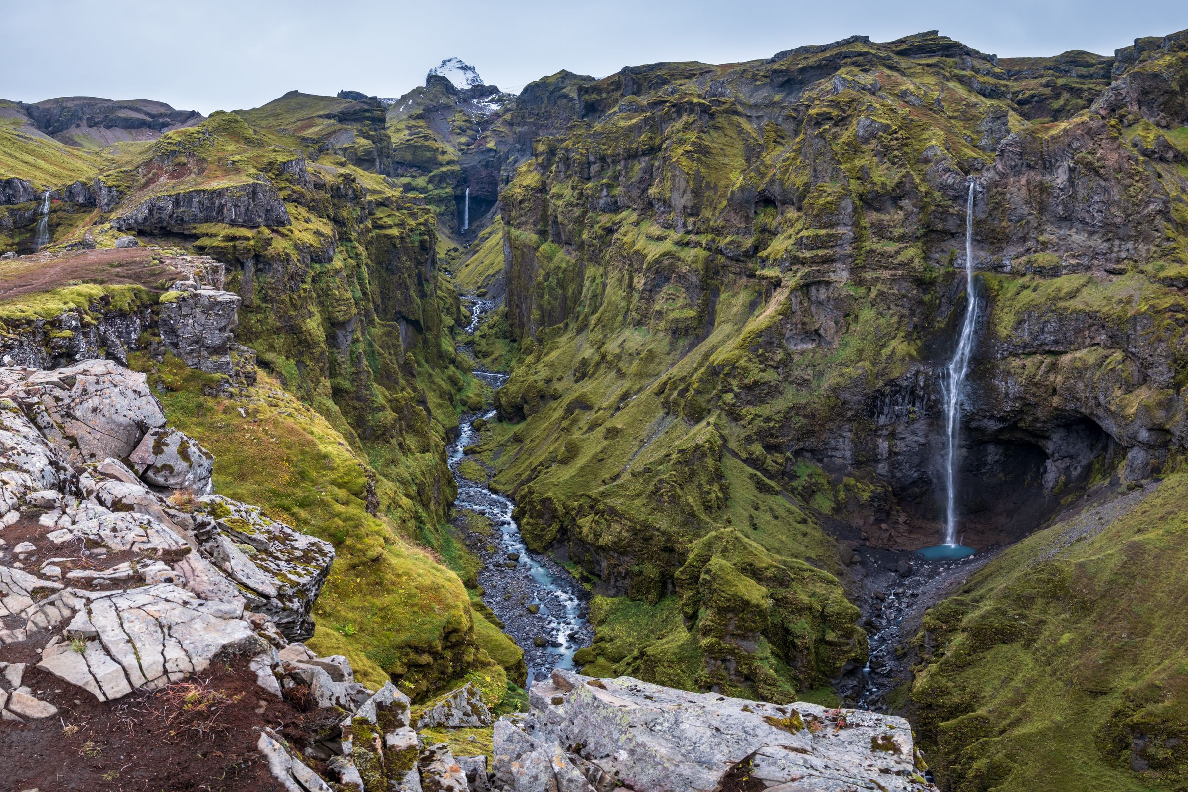 A deep, green canyon with a winding river, multiple waterfalls, and a distant snow-capped mountain.