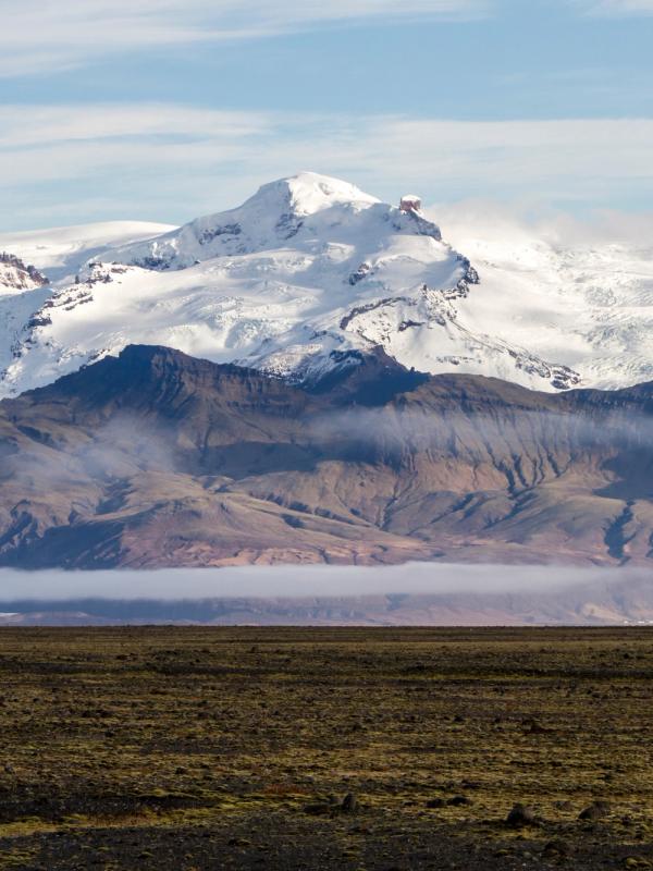 A dark, rocky plain leads to brown mountains veiled in mist, with large snow-capped glaciers in the distance under a blue sky.