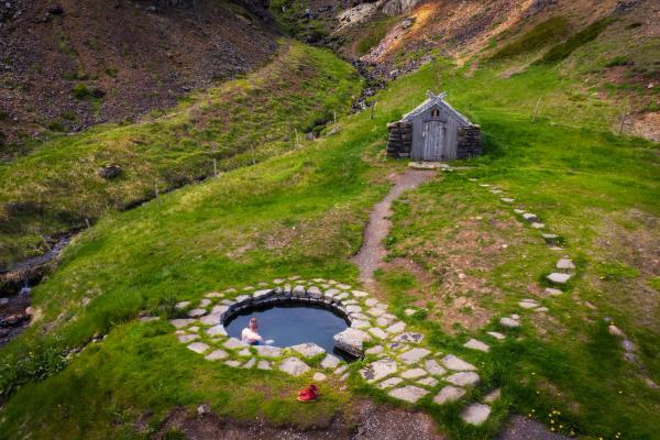 A man soaking in a stone-lined hot spring pool in a green valley, with a rustic hut nearby.