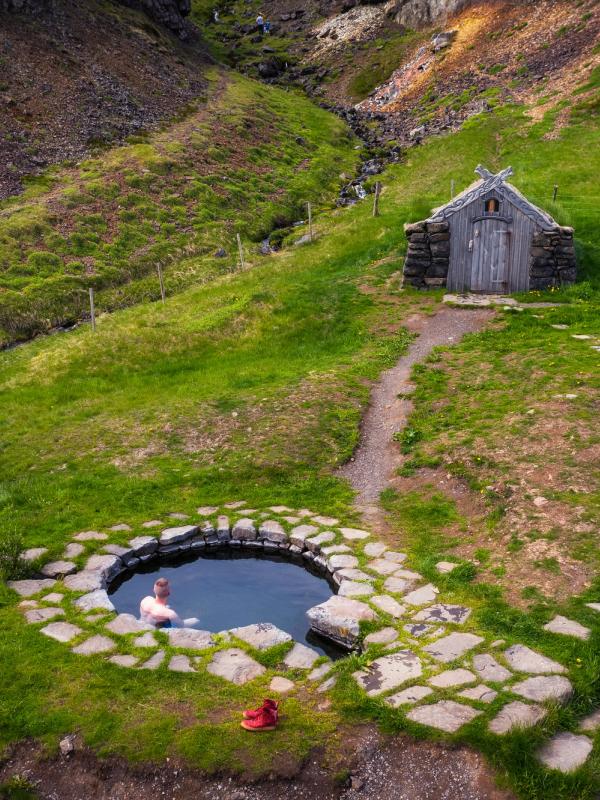 A person relaxes in a stone-lined hot spring on a green hillside, with a small, rustic hut and a path leading up to it.