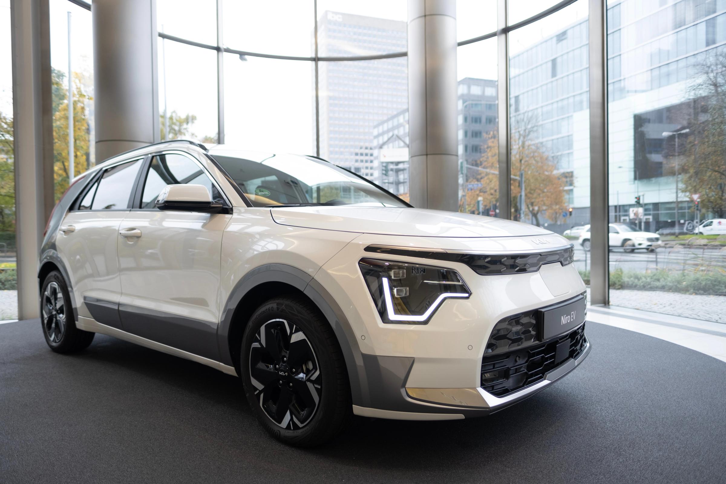 Hvítur Kia Niro EV á hringlaga sýningarpalli í sýningarsal með glerveggjum, séður skáhallt að framan