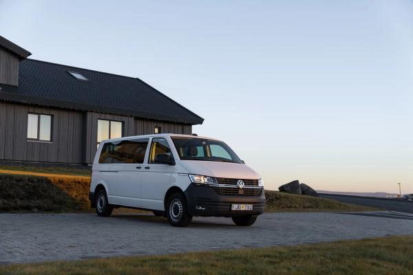 A white Volkswagen van parked on a paved lot in front of a modern house under a clear sky.