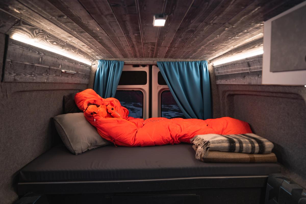 Cama trasera con saco de dormir naranja y manta doblada bajo un techo revestido de madera con iluminación LED