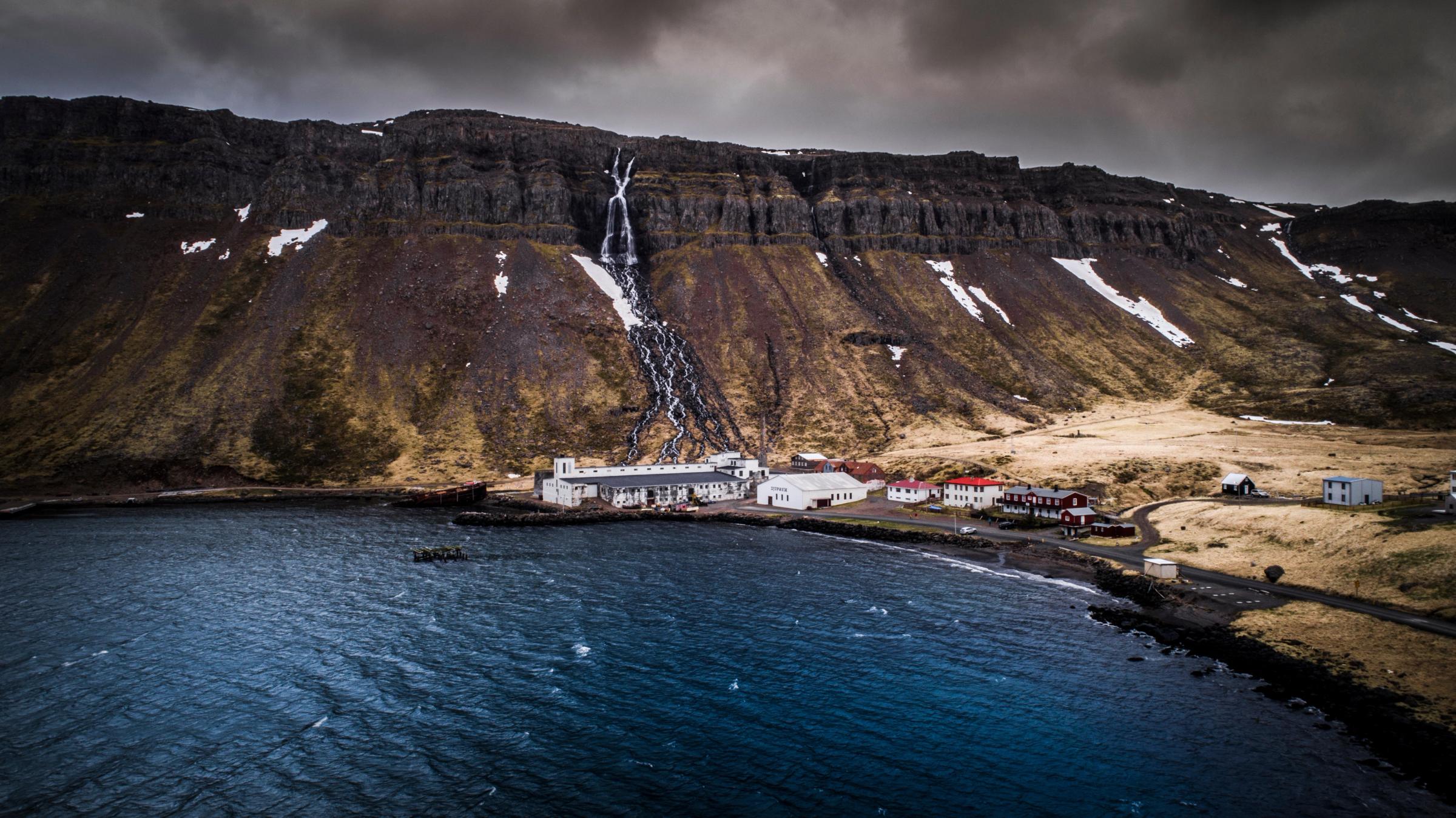 A small coastal town with a waterfall cascading from a large, snow-dusted mountain into a dark bay under a cloudy sky.
