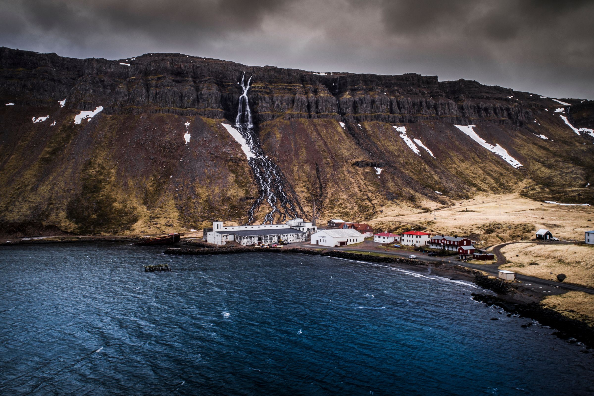 A small coastal town with a waterfall cascading from a large, snow-dusted mountain into a dark bay under a cloudy sky.
