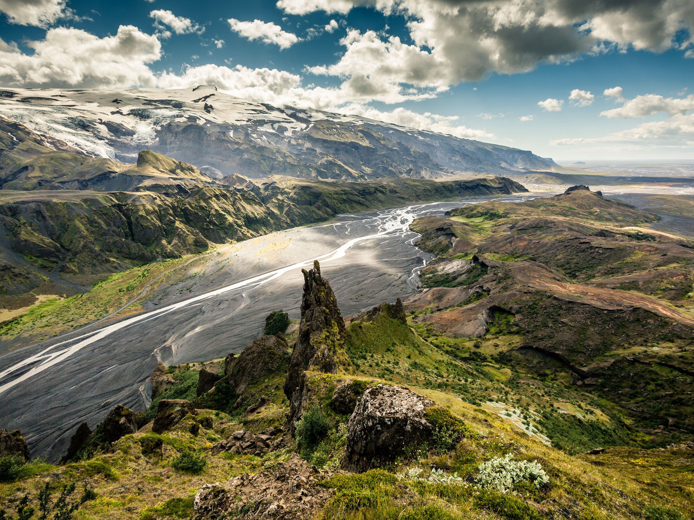 Þórsmörk Dramatic landscape of a braided river valley, with green and rocky terrain, a tall rock spire, and a distant glacier under a cloudy sky.