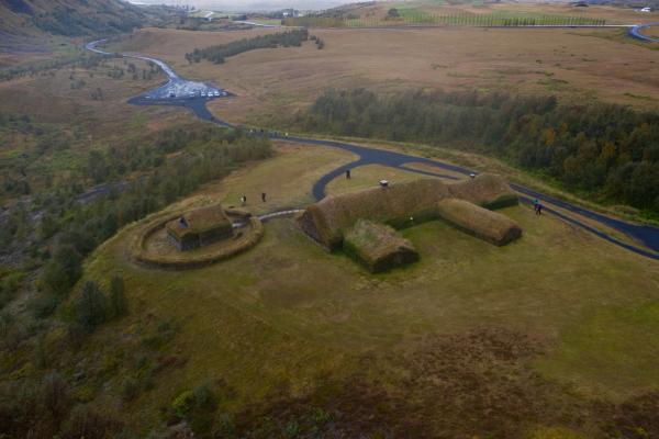 Aerial view of grass-covered turf houses, one within a circular enclosure, set in a vast green landscape with winding paths.