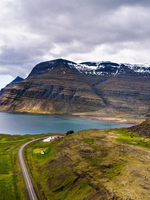 Aerial view of a winding road along a fjord with green hills and snow-capped mountains under a cloudy sky.