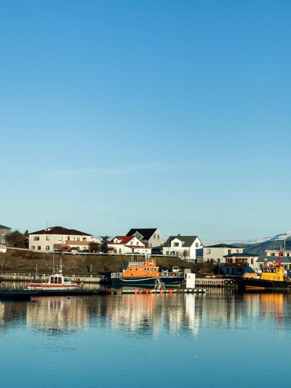 A tranquil harbor town with houses, docked boats, and snow-capped mountains reflecting in calm water under a clear blue sky.