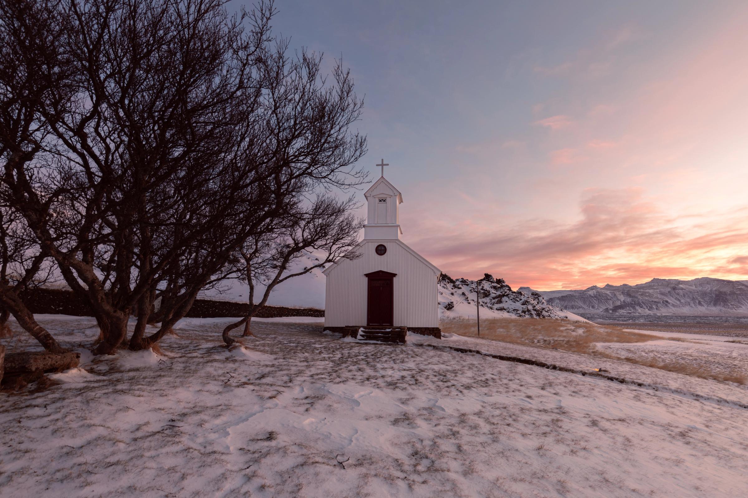White church and bare tree on a snowy hill under a colorful sunset sky.
