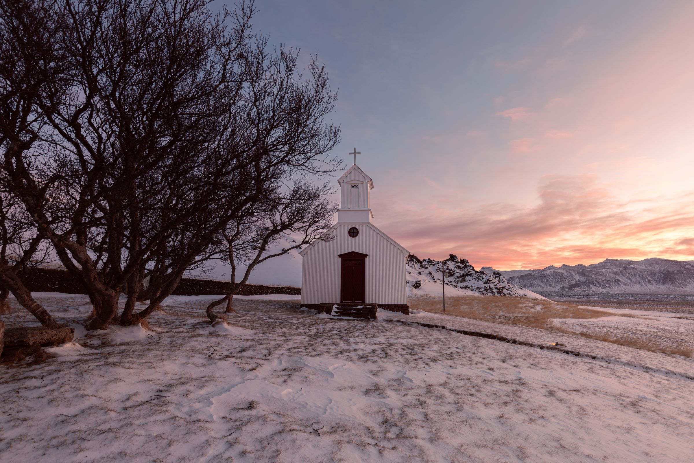 White church and bare tree on a snowy hill under a colorful sunset sky.