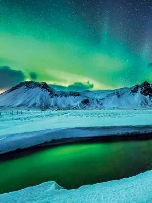 Aurora boreal verde sobre montañas nevadas y un río reflejante.