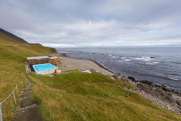 Geothermal pool and buildings on a grassy hillside overlooking a rocky coast and ocean under a cloudy sky.