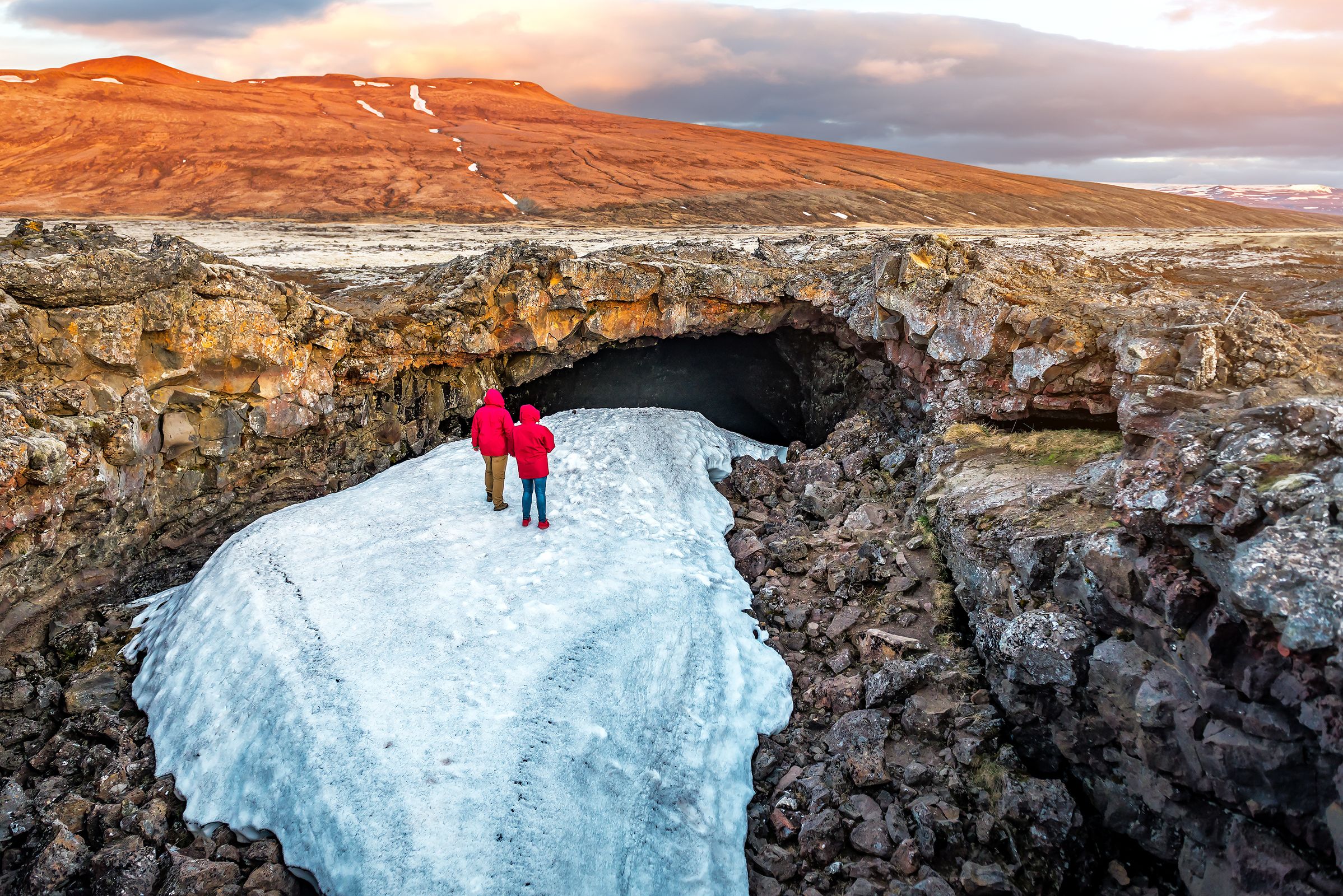 Dos personas con chaquetas rojas caminan sobre la nieve hacia la entrada de una cueva oscura en un paisaje volcánico y accidentado.