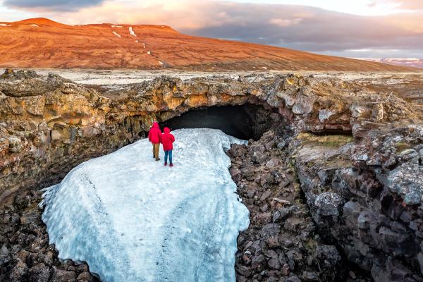 Two people in red jackets walk on snow towards a dark cave entrance in a rugged, volcanic landscape.