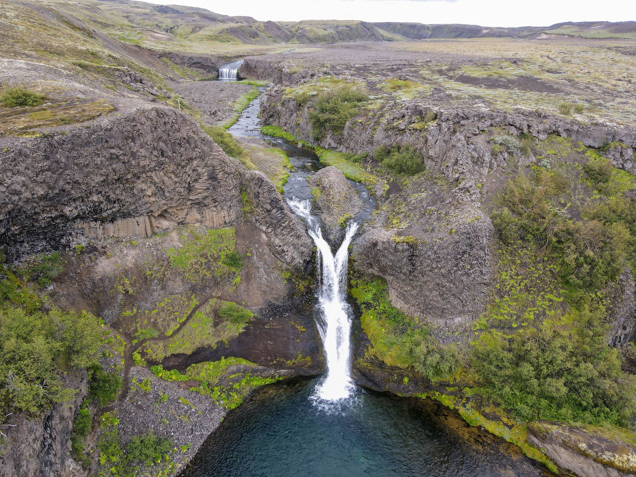 A dual-stream waterfall plunges into a clear blue pool in a rocky, mossy canyon, with a river and distant waterfall.