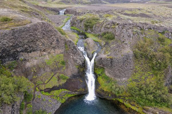 A dual-stream waterfall plunges into a clear blue pool in a rocky, mossy canyon, with a river and distant waterfall.