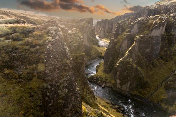 Meandering river through a deep canyon with moss-covered walls under a warm and cloudy sky.