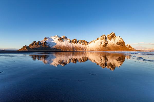 Snow-capped mountains reflected in wet black sand under a clear blue sky.