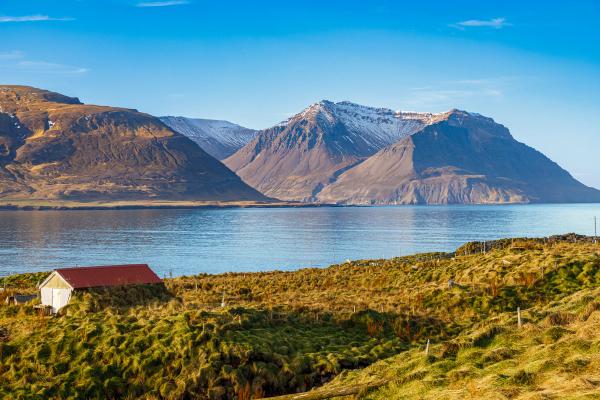a small house with a red roof is sitting on the shore of a lake with mountains in the background .