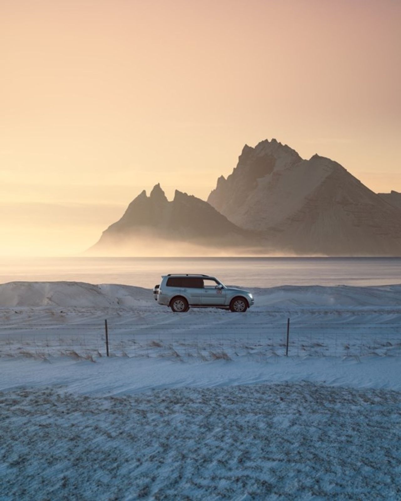 Coche blanco en un prado nevado con montañas y agua en el fondo