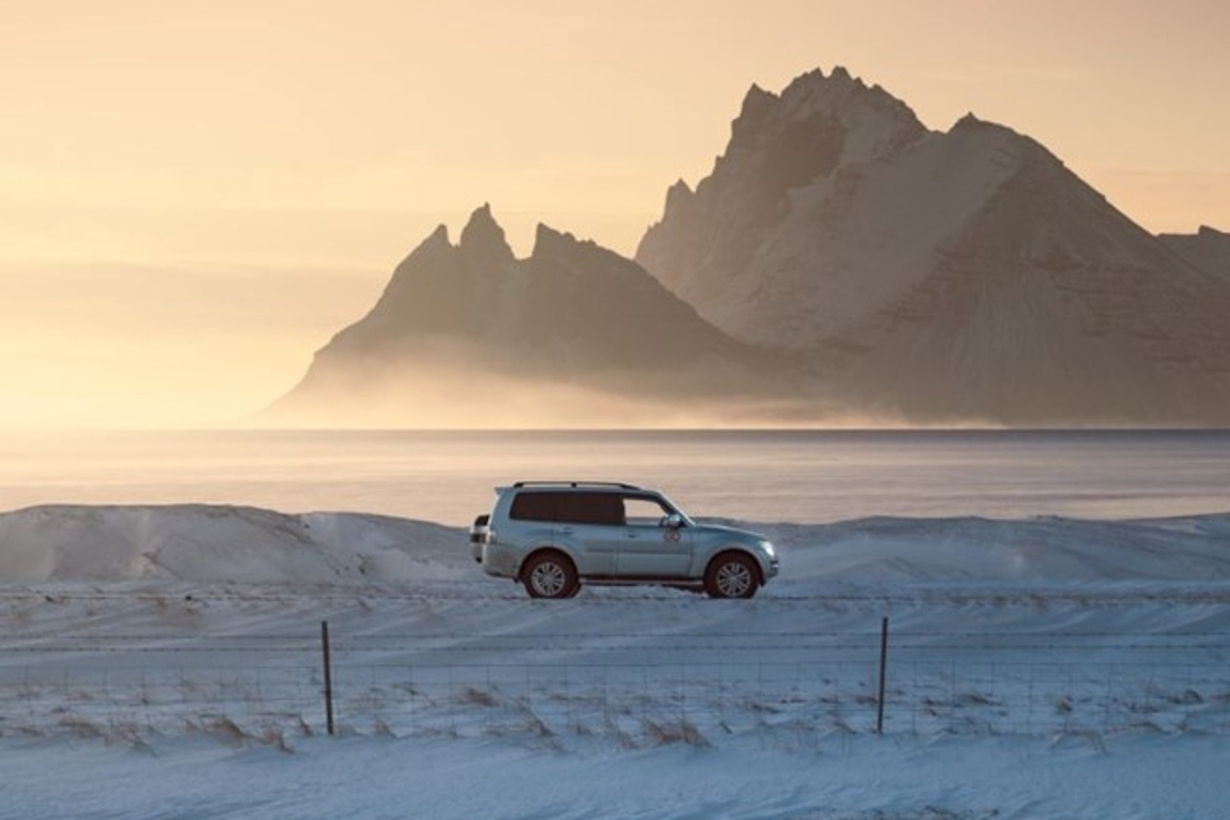 Coche blanco en un prado nevado con montañas y agua en el fondo