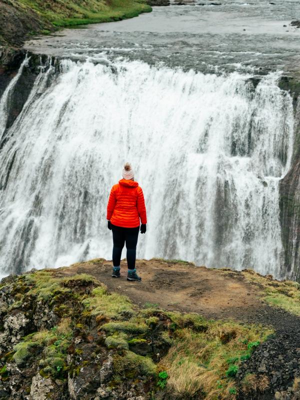A person in an orange jacket and white hat stands on a rocky outcrop, facing a wide waterfall.