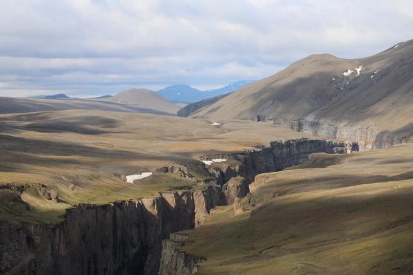 A deep canyon carves through a vast, barren landscape with distant hills and scattered snow patches under a cloudy sky.