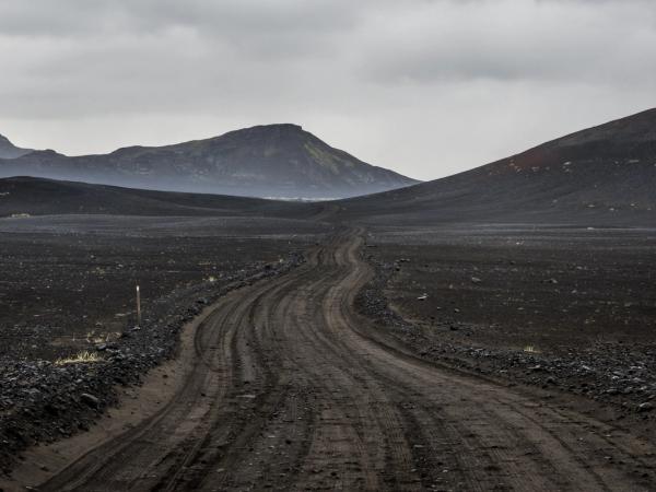 A dirt road winds through a barren, dark volcanic landscape under a cloudy sky.