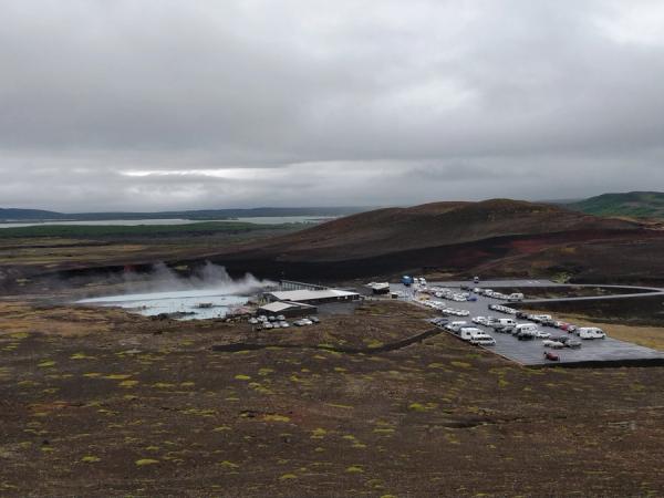 Vista aérea de un balneario geotérmico con aguas azules humeantes, un estacionamiento lleno de vehículos y colinas áridas bajo un cielo nublado.