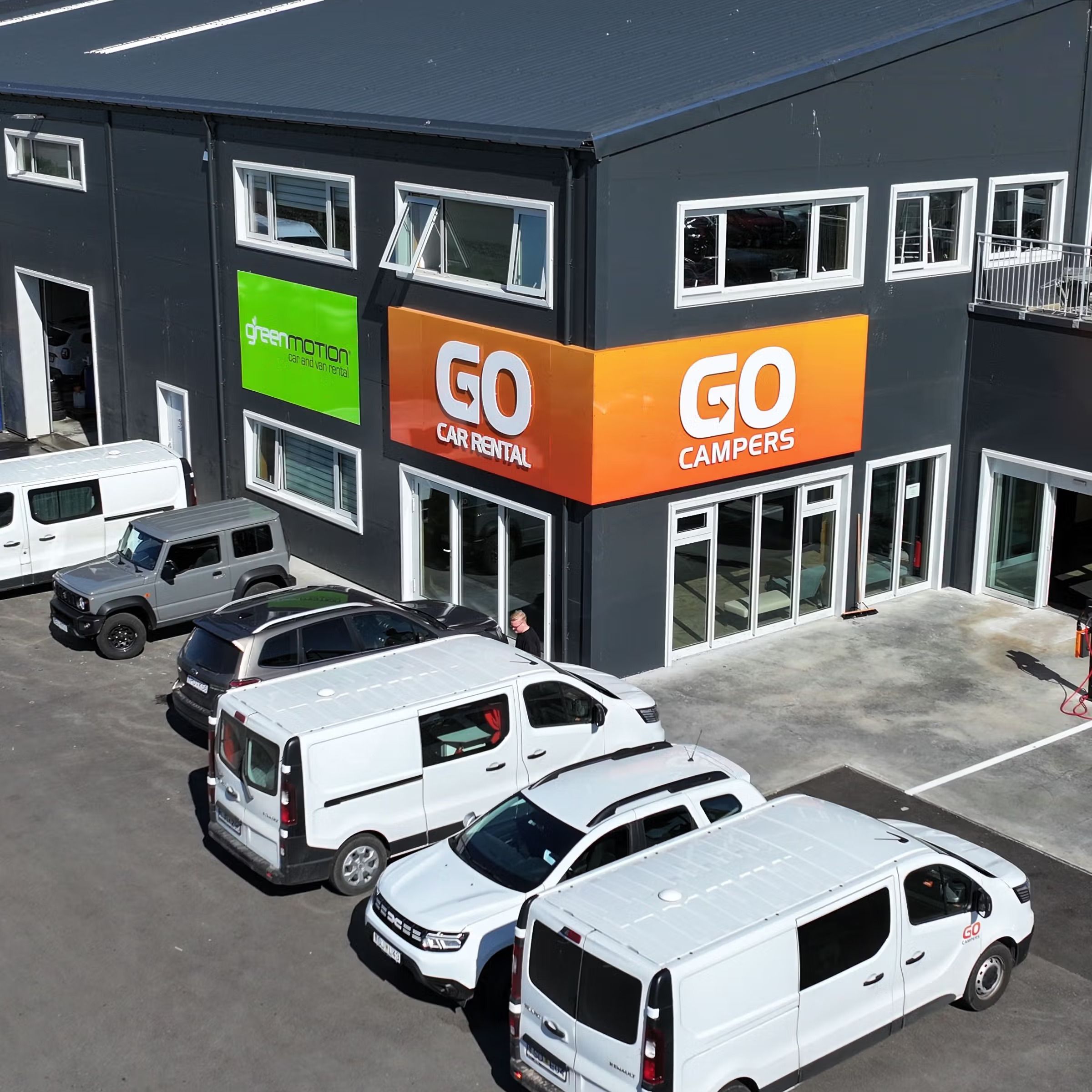 Aerial view of a dark building with "Green Motion", "GO Car Rental", and "GO Campers" signs, with several white rental vans and cars parked outside.