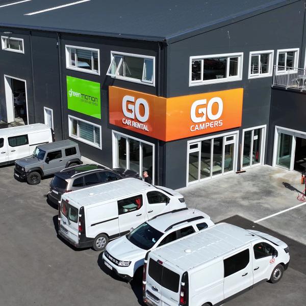 Aerial view of a dark building with "Green Motion", "GO Car Rental", and "GO Campers" signs, with several white rental vans and cars parked outside.