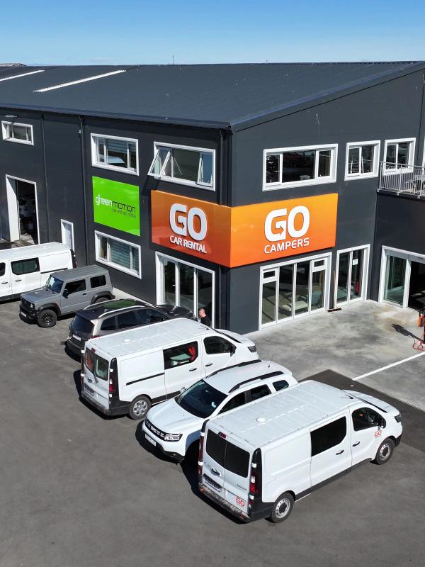 A dark grey building featuring GO Car Rental, GO Campers, and Green Motion signs, with a parking lot full of white vans and cars.